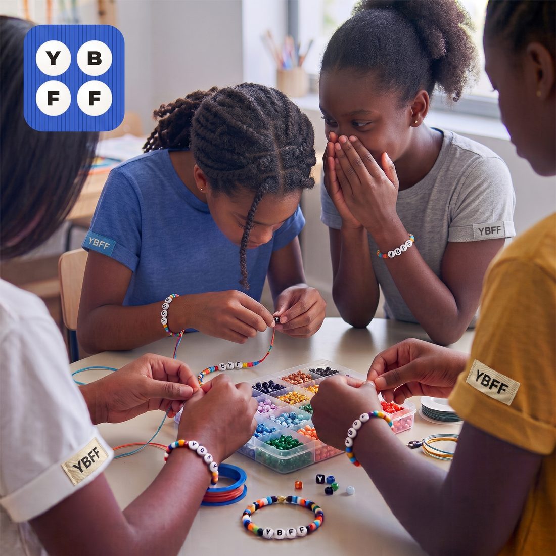 Four girls around a table making beaded friendship bracelets together, one laughing behind her hands. The bracelets and their sleeve labels read YBFF.